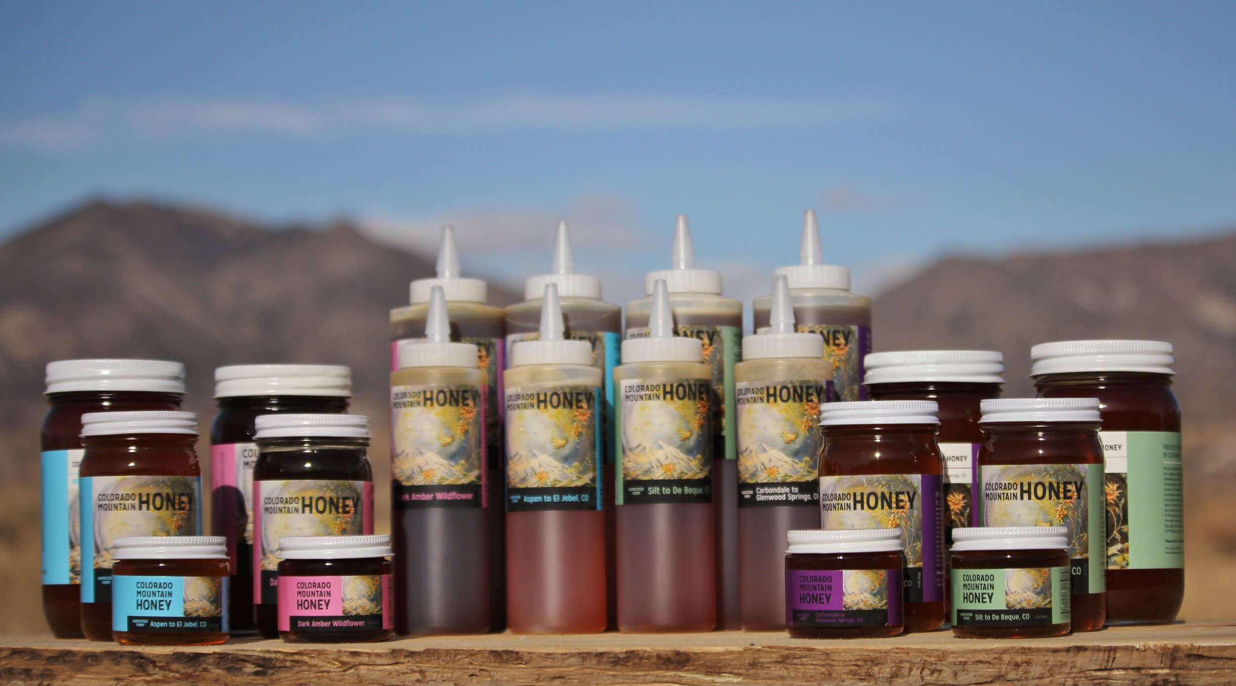 A variety of Colorado Mountain Honey products displayed outdoors, including jars and squeeze bottles, against a scenic mountain backdrop. The image showcases different flavors and sizes, emphasizing the natural and artisanal quality of the honey.