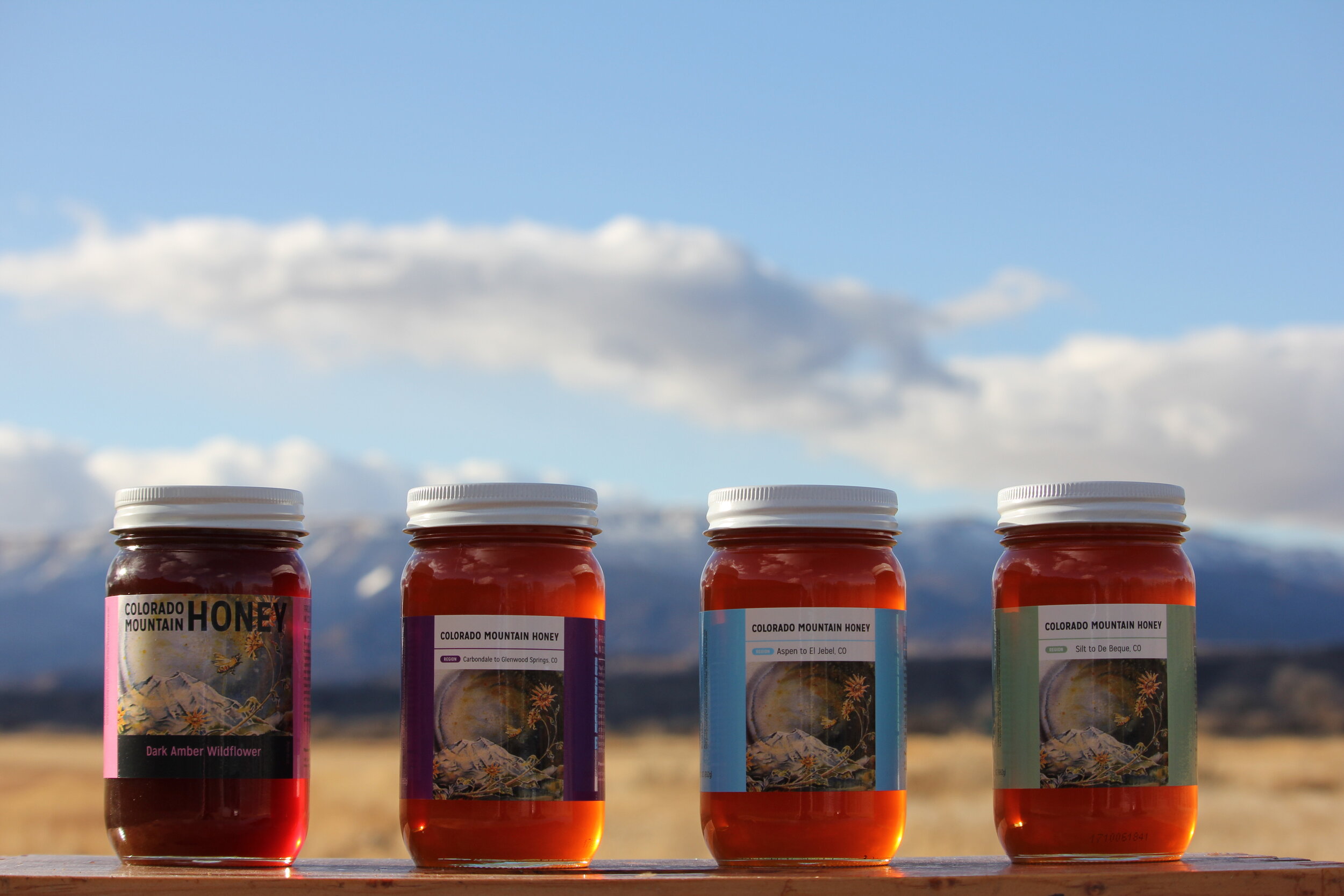 Jars of Colorado Mountain Honey displayed outdoors against a backdrop of mountains and blue sky, showcasing different varieties including Dark Amber Wildflower, Aspen to Jebel, and Silt to De Beque.